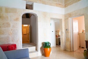 A stone-walled interior with a staircase leading to a second floor, a large orange vase, and a red suitcase on a blue sofa.