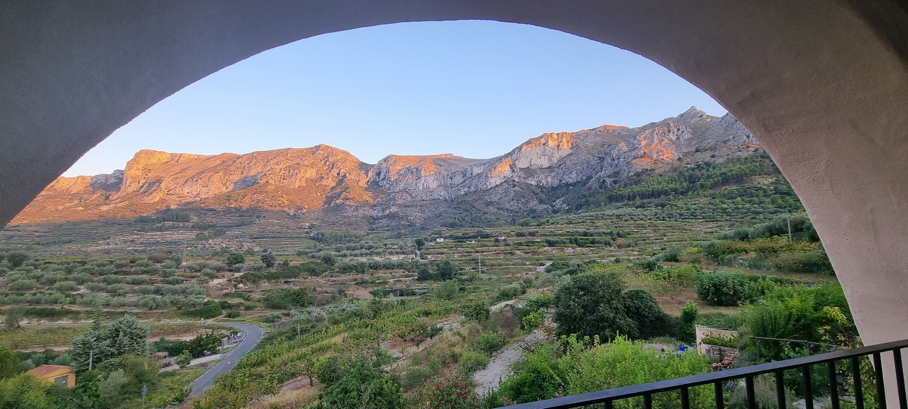 Paesaggio montuoso visibile attraverso una finestra ad arco, con una strada e vegetazione in primo piano.