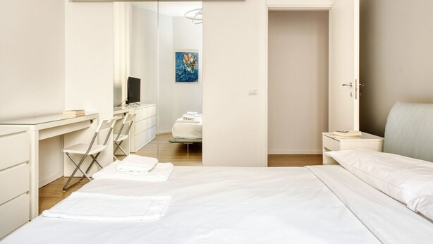 A modern hotel room with a bed, a desk, a chair, a small table, and a door leading to another room. - Hostmate - Milano - Donatello - Città Studi (Milano)