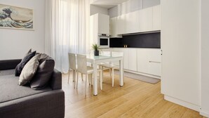 A modern living room with a sofa, dining table, and chairs, a kitchen with white cabinets and a black countertop, and a painting on the wall. - Hostmate - Milano - Donatello - Città Studi (Milano)
