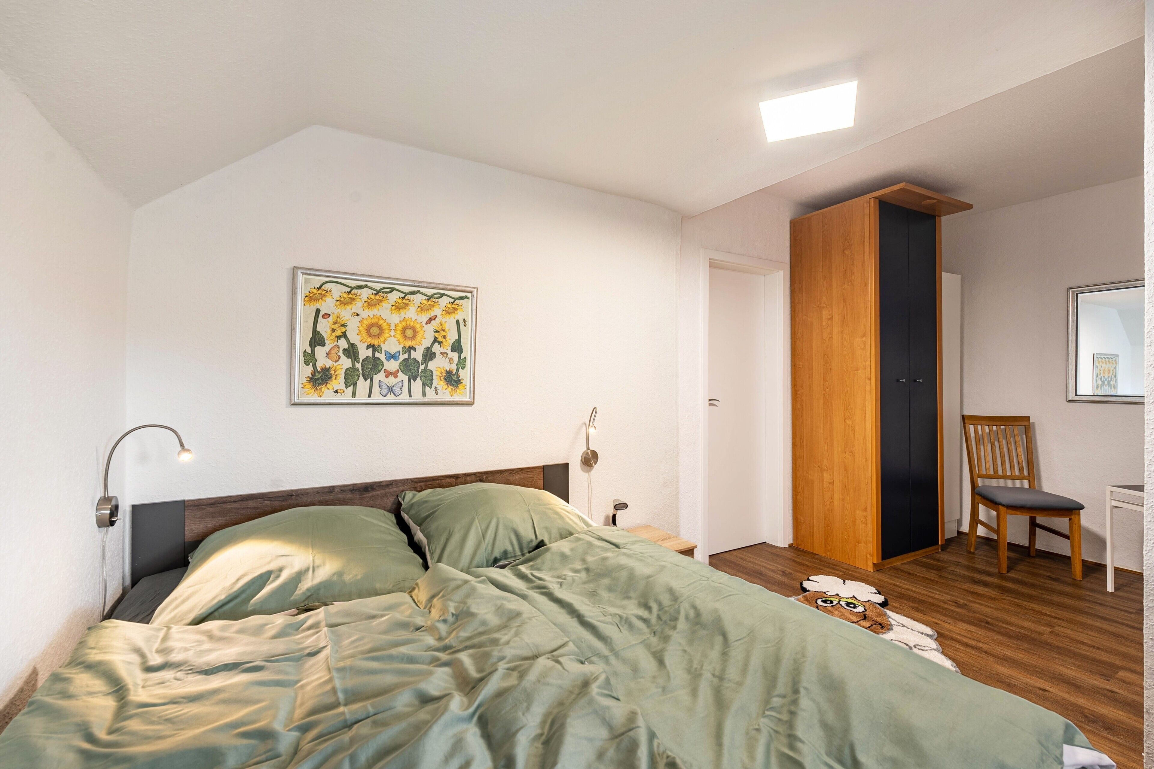 A bedroom with a bed, a wall-mounted lamp, a painting of flowers, a wooden wardrobe, and a chair.
