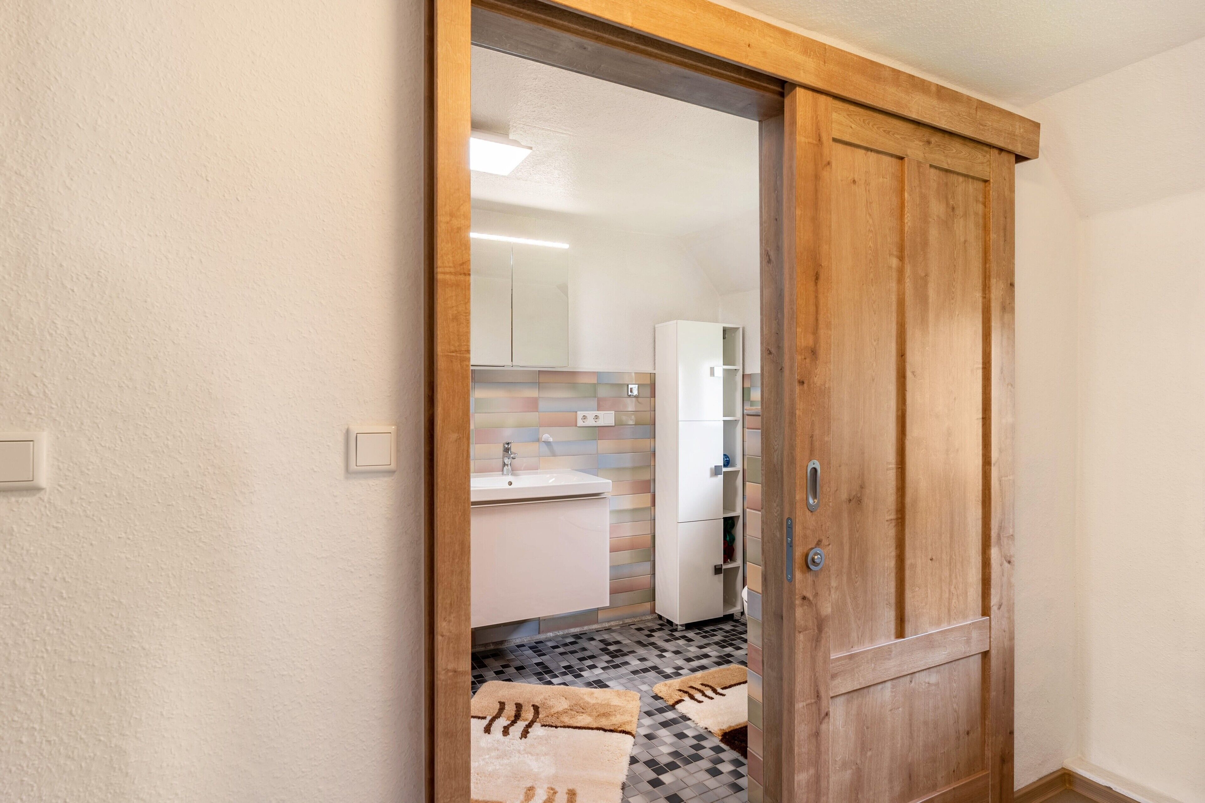 Een houten deur die leidt naar een badkamer met betegelde vloer en een spiegel.