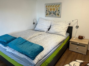 A neatly made bed with patterned bedding, a wall-mounted artwork, a bedside table with a kettle, and a nightstand with a camera. - Holiday Home 'Reethaus Kälberstieg' with Lake View, Private Terrace and Wi-Fi (Grebin)