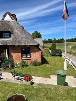 A thatched-roof house with a brick base, a flagpole with a flag, a bench, a flower pot, and a green trash bin. - Holiday Home 'Reethaus Kälberstieg' with Lake View, Private Terrace and Wi-Fi (Grebin)