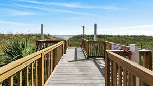 Een houten looppad dat naar een strand leidt, met een bord waarop staat dat je jezelf moet afspoelen voordat je het strand betreedt.