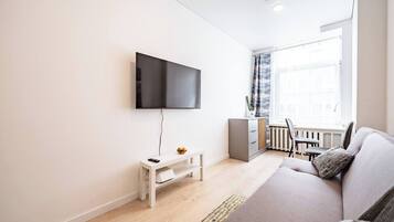 A modern living room with a flat-screen TV mounted on the wall, a grey sofa with a throw pillow, a small white side table with a remote control, and a dining area with a table and chairs.
