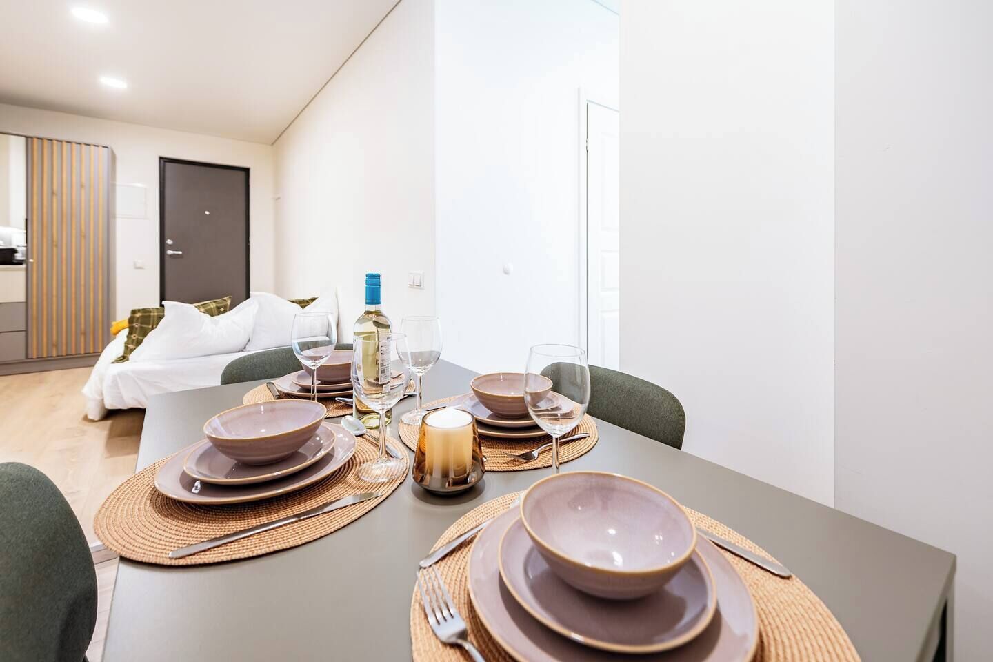 Une table à manger dressée avec des assiettes, des bols, des couverts et une bougie, dans une pièce où se trouvent un lit et une armoire.