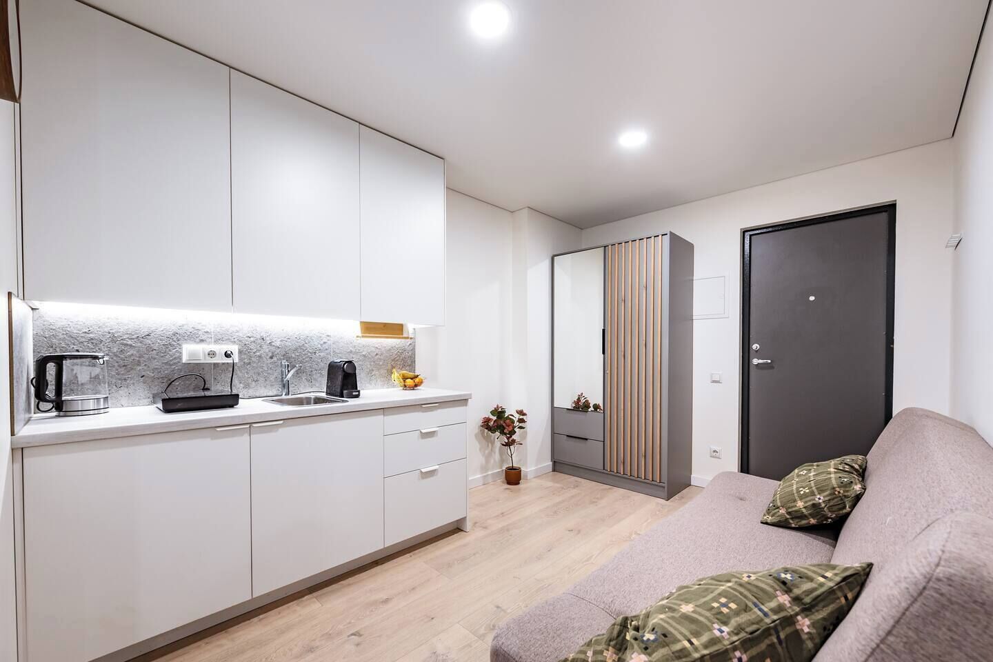 A modern kitchen with white cabinets, a countertop, and a kettle. There is a sofa with patterned cushions and a door in the background.