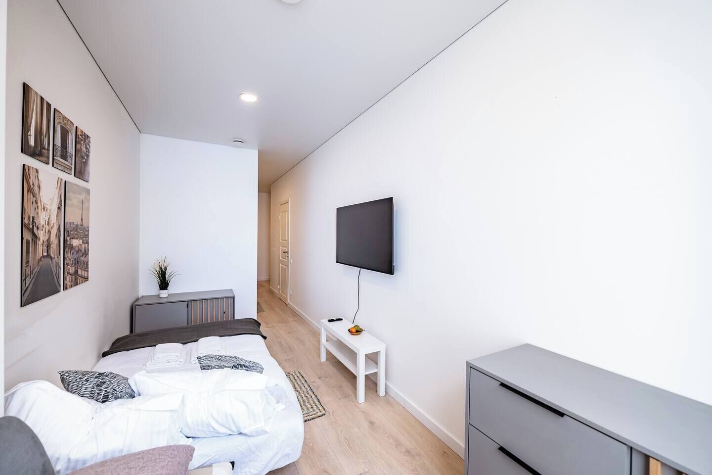 A modern bedroom with a bed, a flat-screen TV mounted on the wall, and a small white table with a plant.