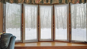 A bay window with wooden frames and white blinds, overlooking a snowy outdoor scene. - Lake Access w/ Dock! Hayward Cabin Hideaway (Hayward)
