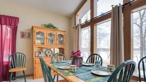 A dining room with a wooden table set for four, surrounded by green chairs. A wooden cabinet with glass doors is against the wall, and a window with curtains is visible. - Lake Access w/ Dock! Hayward Cabin Hideaway (Hayward)