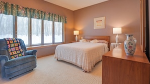 A bedroom with a bed, two bedside lamps, a wooden nightstand, a blue armchair with a colorful blanket, a decorative vase, and a ceiling fan. - Lake Access w/ Dock! Hayward Cabin Hideaway (Hayward)