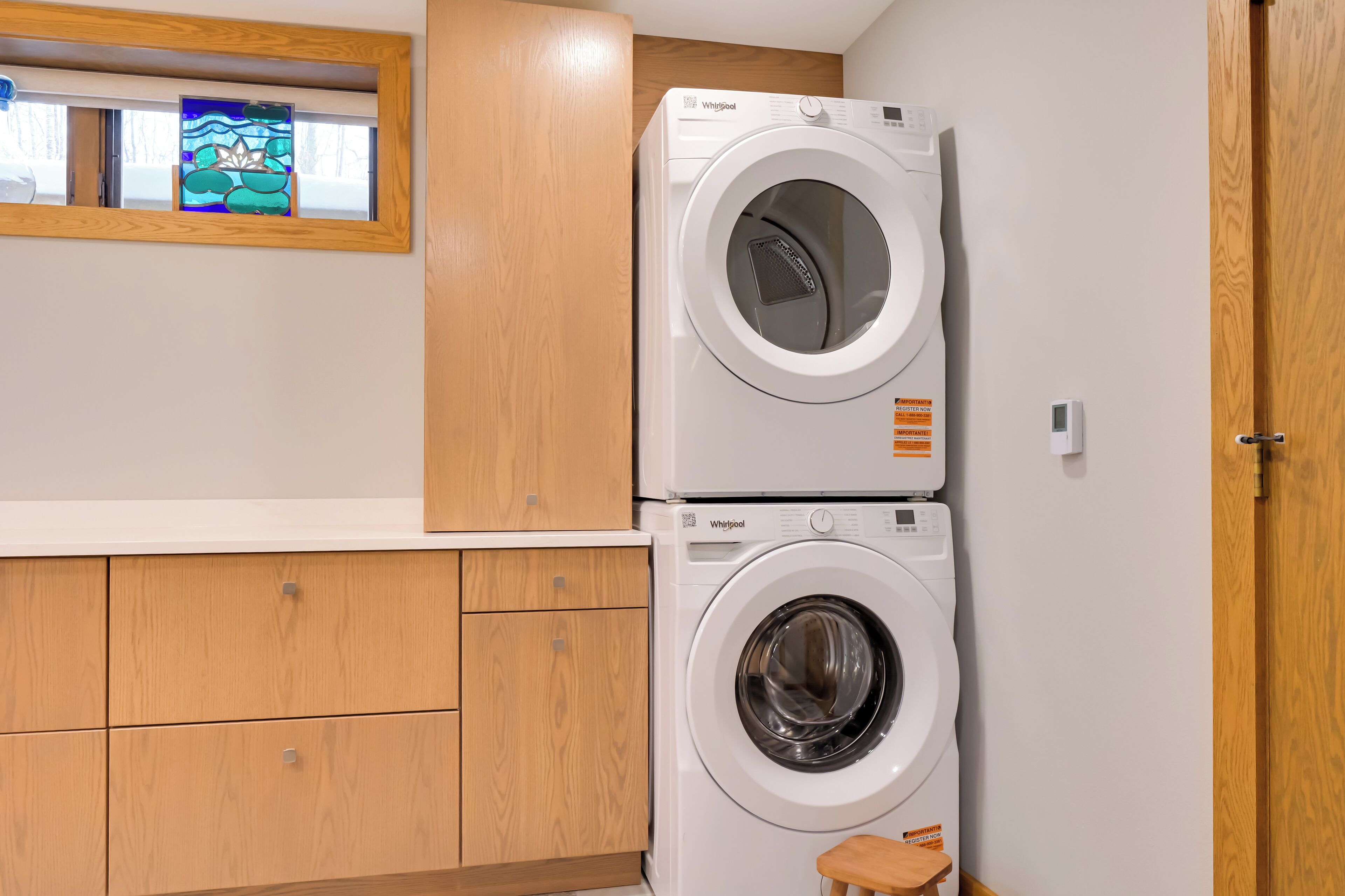 Un lavadero con dos lavadoras Whirlpool apiladas, armarios de madera y un ventanal con diseño de vitral.