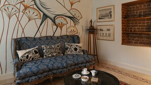 Een kamer met een blauwe bank, een ronde salontafel, een kroonluchter en een muurschildering van een vogel en bloemen.
