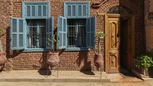 Een gebouw van baksteen met twee openslaande blauwe luiken, een houten deur en potplanten.