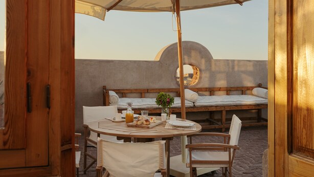 Een terras met een houten tafel en stoelen, een grote parasol en een bed met een rond raam.