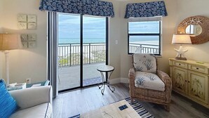 Een kamer met uitzicht op zee, een rieten stoel, een klein tafeltje en een kast met decoratieve objecten.