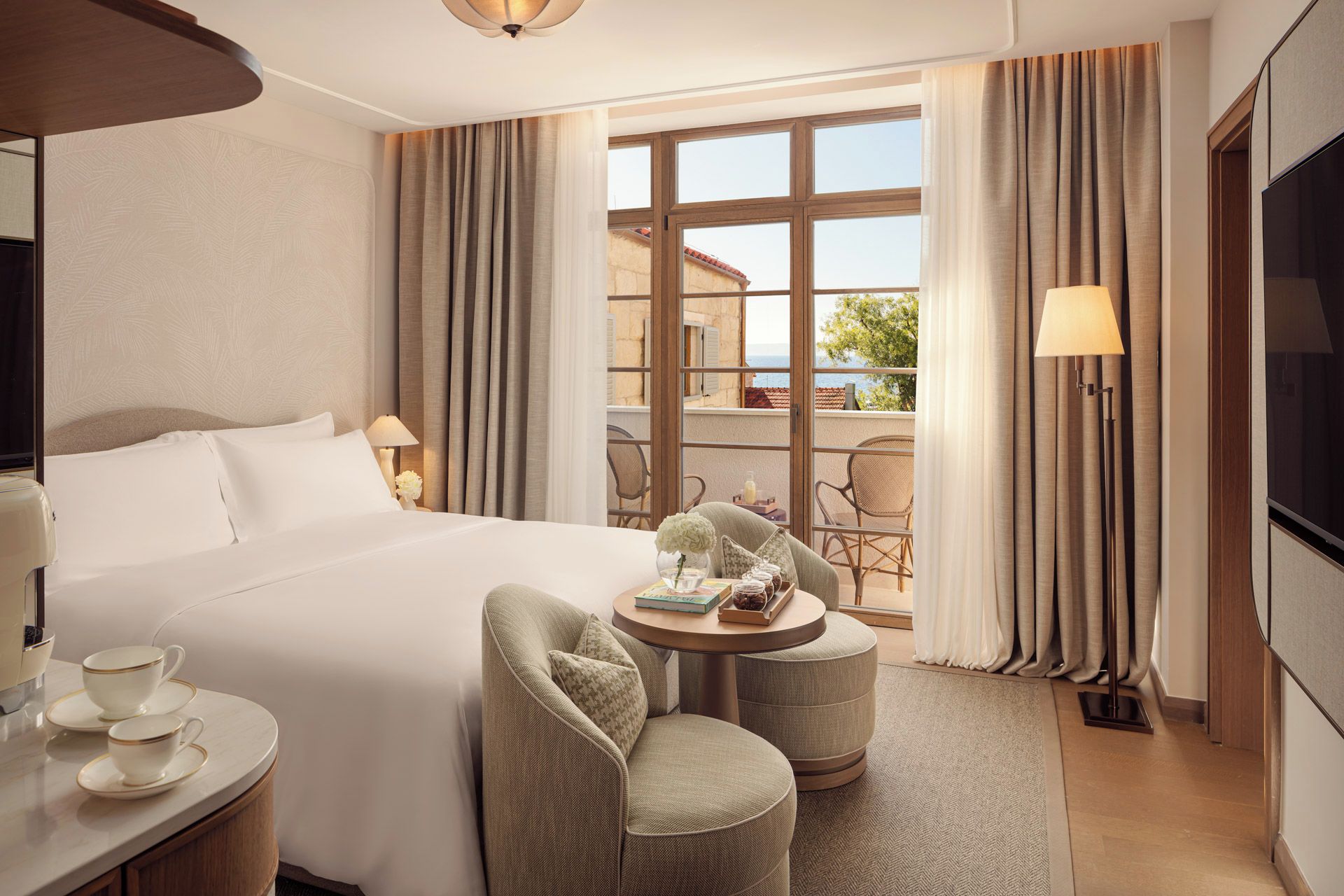 Habitación de hotel con una cama grande, una mesa redonda con sillas, un televisor y vistas al mar a través de amplias ventanas.