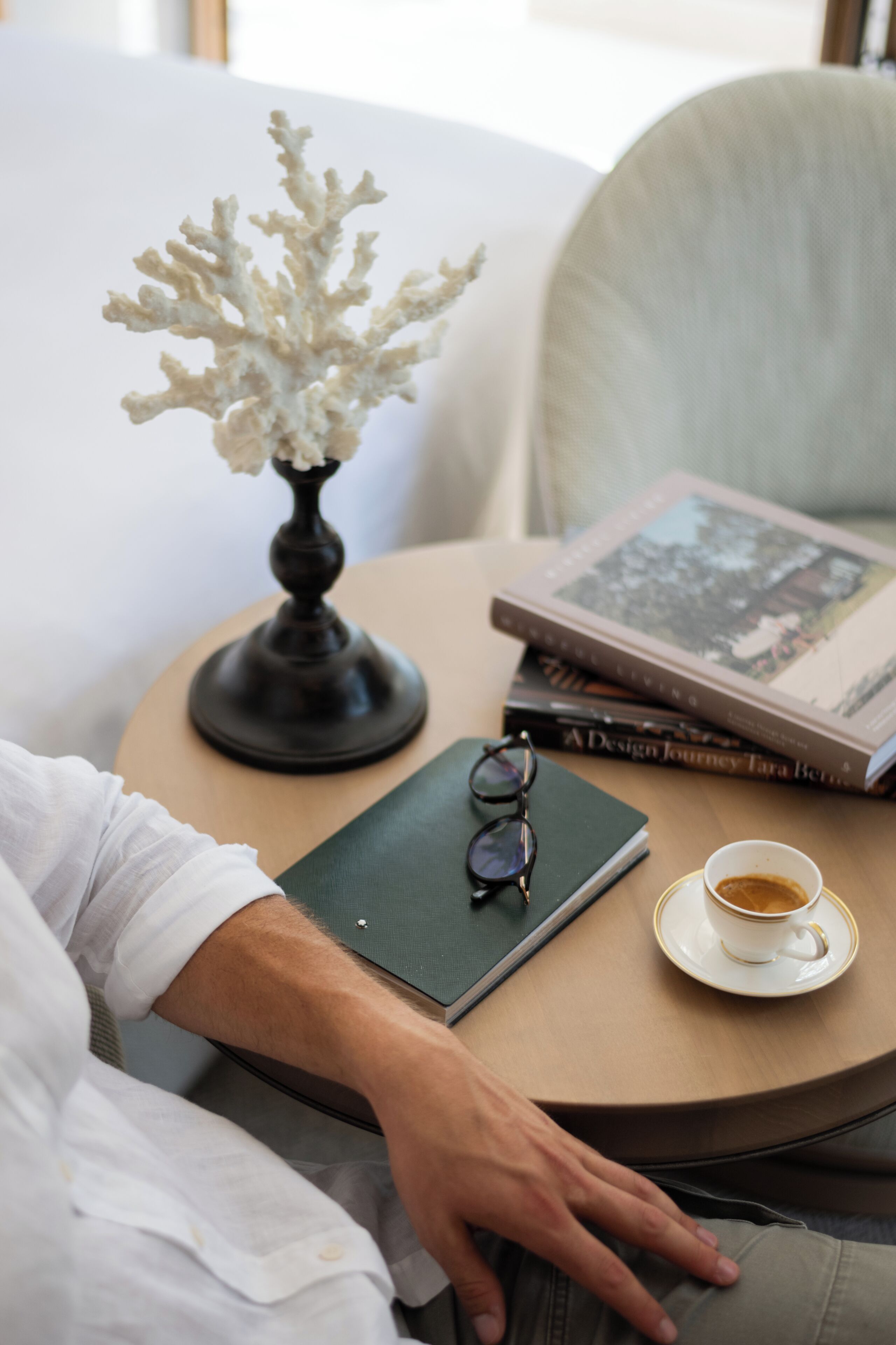 A person seated at a round table with a book, a pair of glasses, and a cup of coffee.