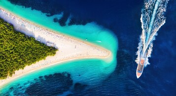 An aerial view of a beach with a boat wake, clear turquoise waters, and a dense forested area.