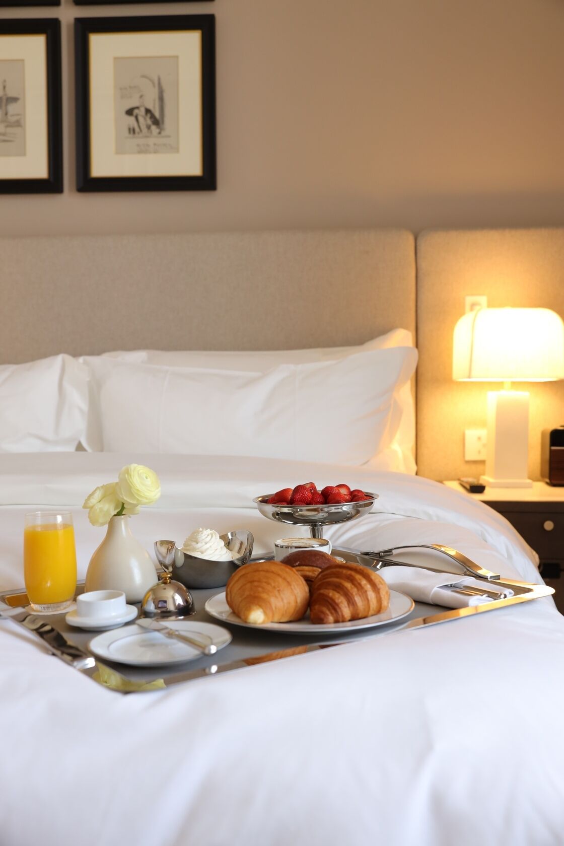 A neatly made bed with white linens, a bedside table with a lamp, and a breakfast tray with croissants, strawberries, and a glass of orange juice.