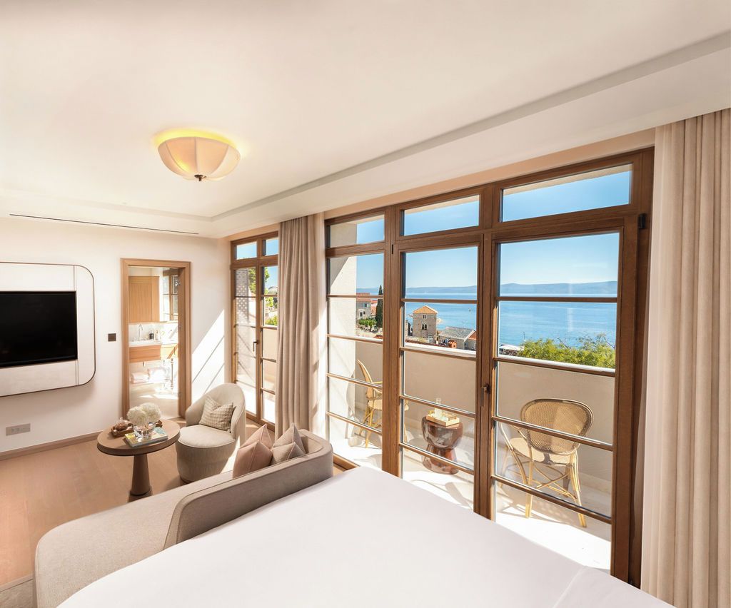 A hotel room with a large bed, a TV, a balcony with a view of the sea, and a dining area with chairs.