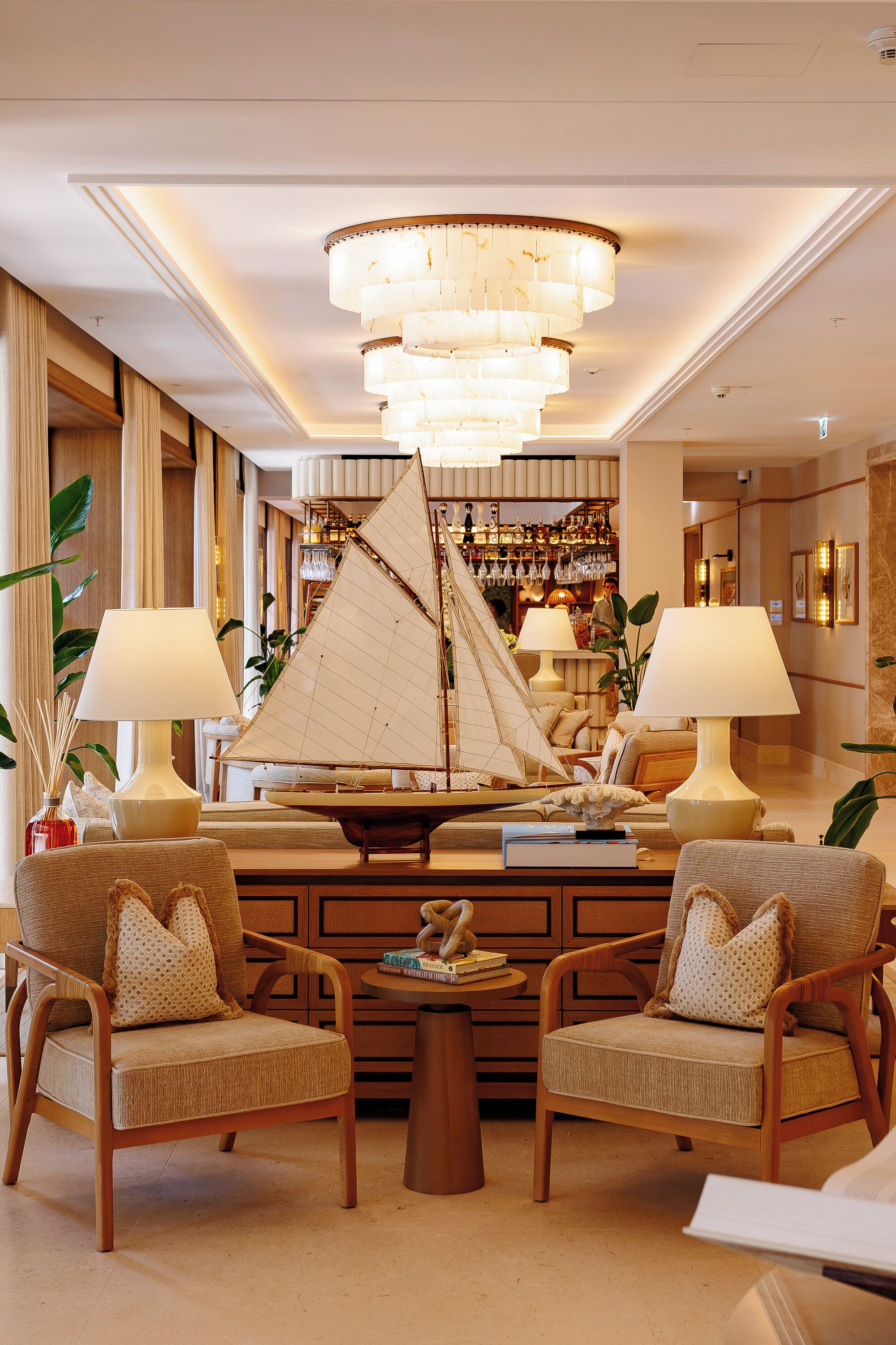 A well-lit lobby with a large chandelier, two armchairs, a central coffee table, and a sailboat model.