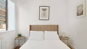 A bedroom with a bed, a bedside table, a lamp, a window, and a framed picture on the wall. - Stylish Feltham Studio near Station & Shops (Vilnius)