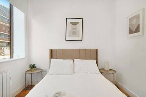Ein Schlafzimmer mit einem Bett, einem Nachttisch, einer Lampe, einem Fenster und einem gerahmten Bild an der Wand.