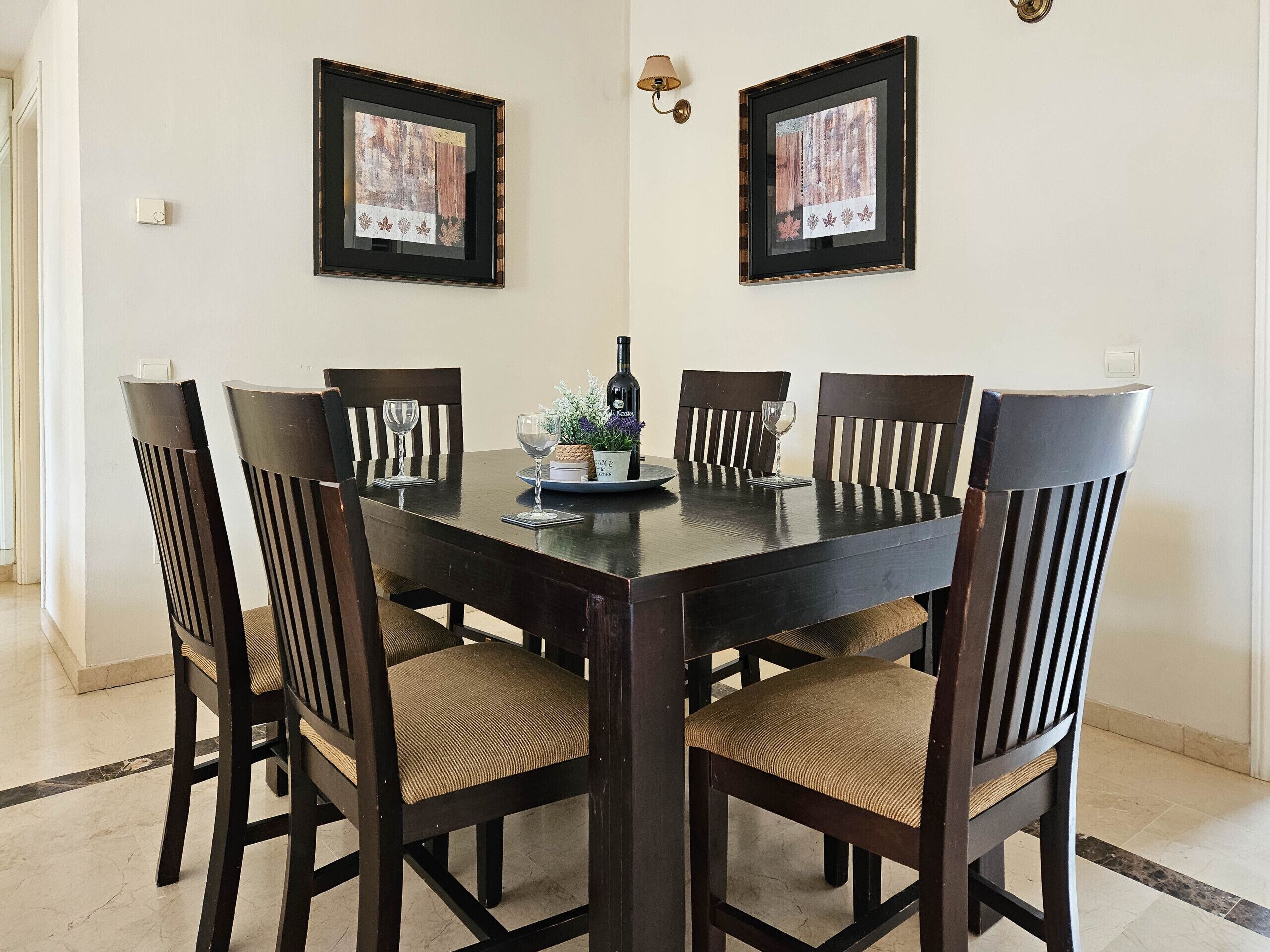 A dining area with a rectangular table surrounded by six chairs, each with a woven seat cushion. Above the table, there are two framed artworks on the wall.