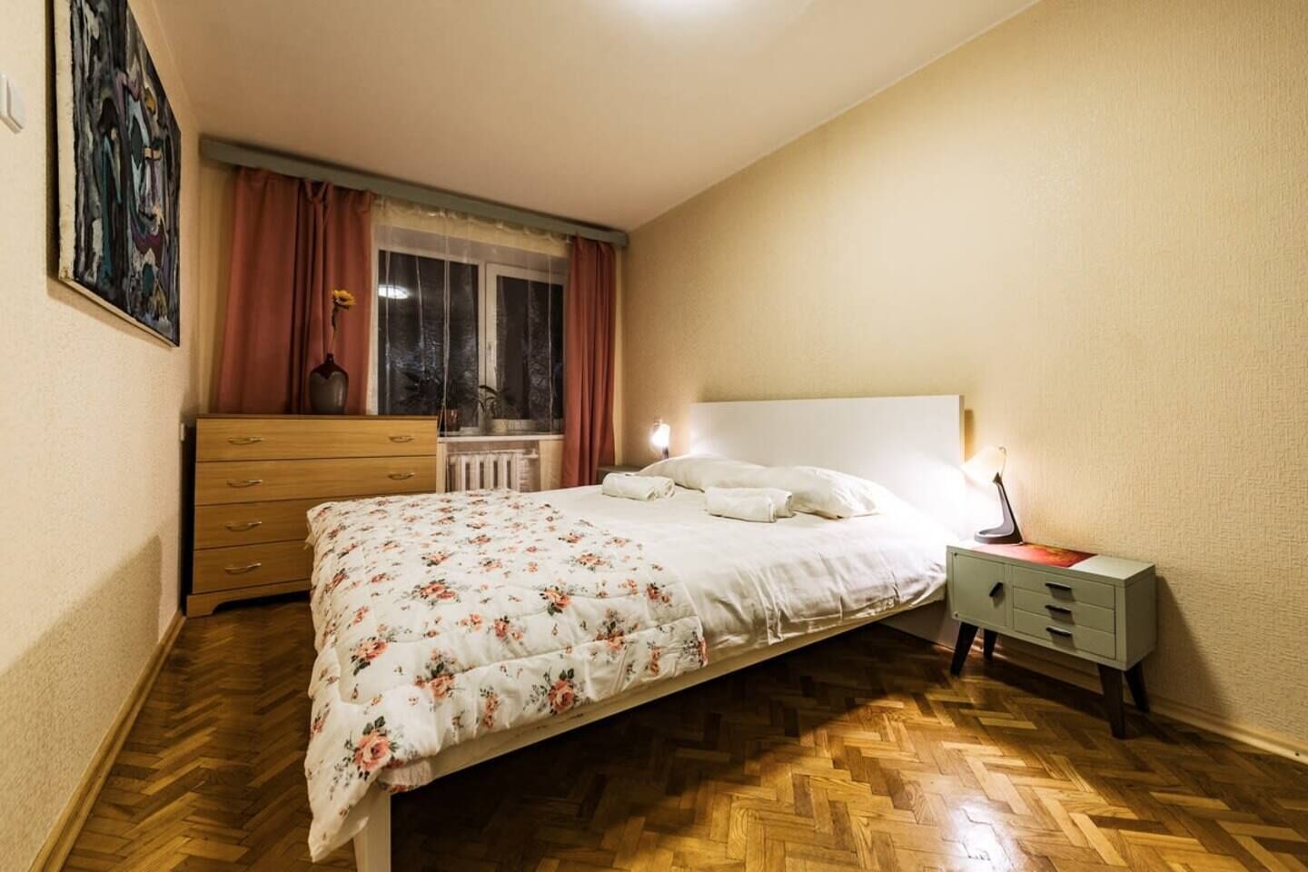 Un dormitorio con cama, cómoda, mesita de noche, ventana con cortinas y radiador.