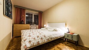 A bedroom with a bed, a dresser, a nightstand, a window with curtains, and a radiator.