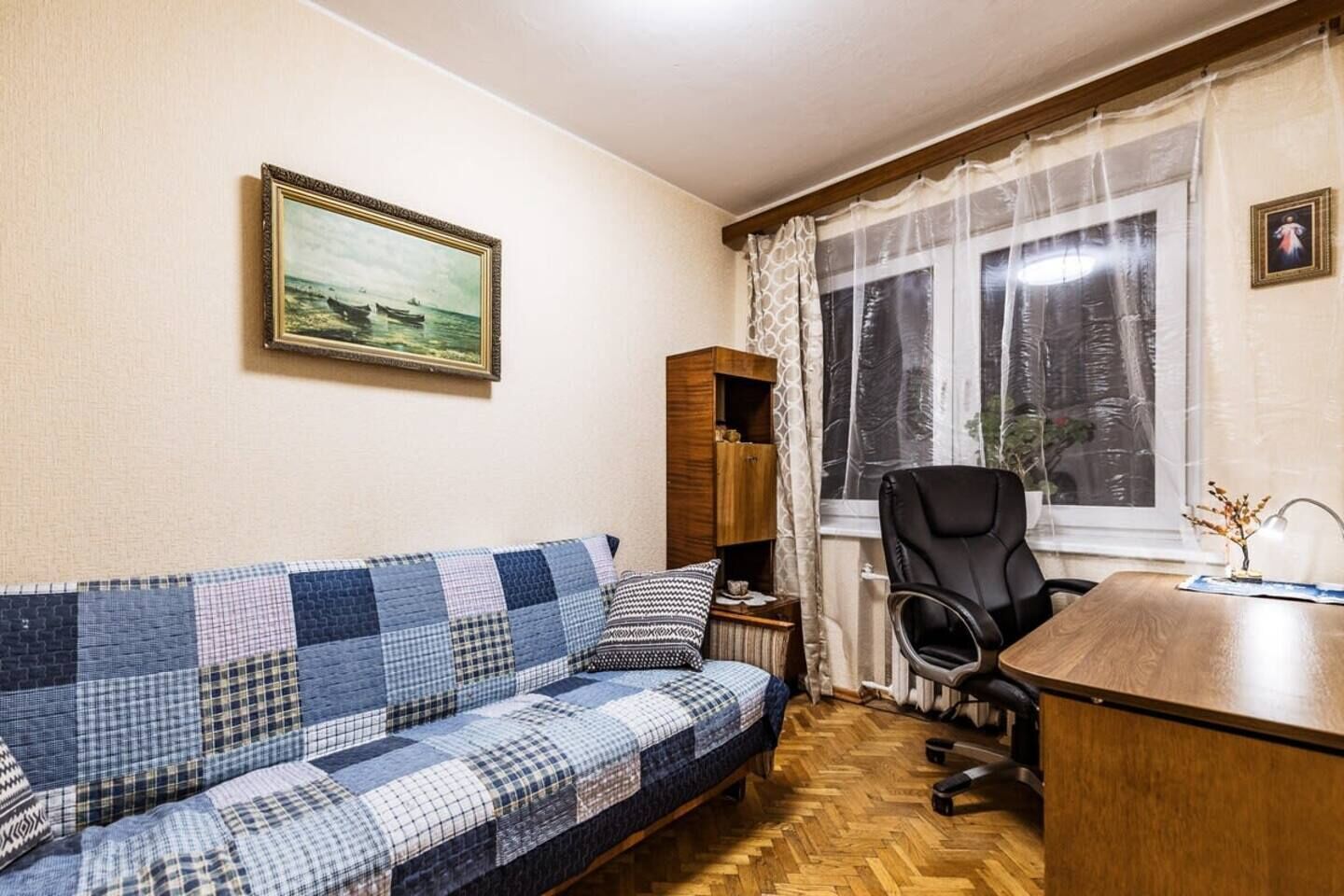 Una habitación con un sofá azul y blanco a cuadros, un escritorio de madera, una silla de oficina negra, una ventana con cortinas translúcidas y un cuadro enmarcado en la pared.