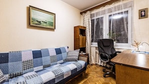 A room with a blue and white checkered sofa, a wooden desk, a black office chair, a window with sheer curtains, and a framed picture on the wall.