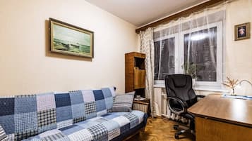 Una habitación con un sofá azul y blanco a cuadros, un escritorio de madera, una silla de oficina negra, una ventana con cortinas translúcidas y un cuadro enmarcado en la pared.