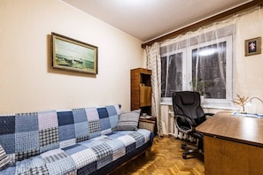 A room with a blue and white checkered sofa, a wooden desk, a black office chair, a window with sheer curtains, and a framed picture on the wall. - Cozy Apartment in Kaunas with Free Parking. (Kaunas)