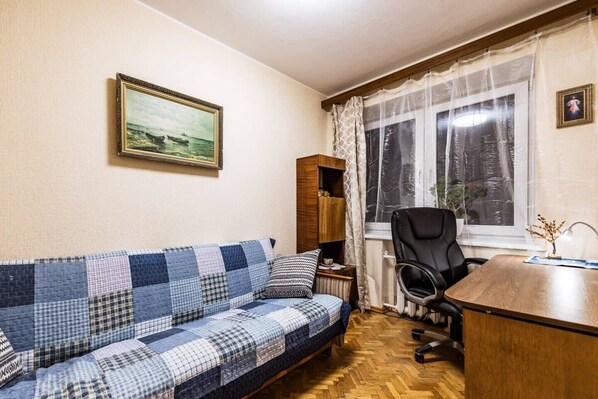 Una habitación con un sofá azul y blanco a cuadros, un escritorio de madera, una silla de oficina negra, una ventana con cortinas translúcidas y un cuadro enmarcado en la pared.