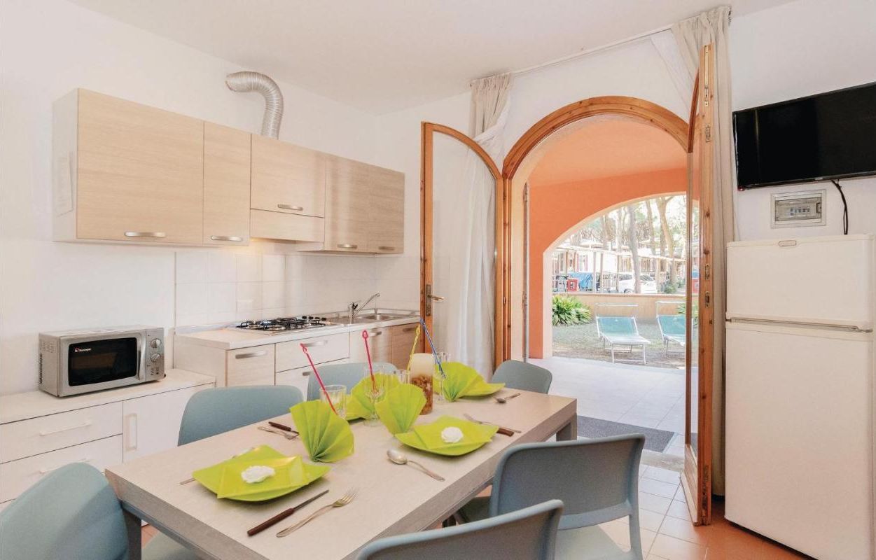A compact kitchen with a microwave, stove, and cabinets. A dining table is set with green placemats and utensils. An open door reveals a pool area with chairs.