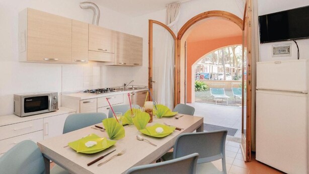 A compact kitchen with a microwave, stove, and cabinets. A dining table is set with green placemats and utensils. An open door reveals a pool area with chairs. - The Charming Marina Village Tipo3-3 Bedroom Villa 6 Persons (Comacchio)