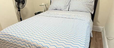 A bed with a patterned bedspread, a bedside table, a fan, and a wall-mounted heater.