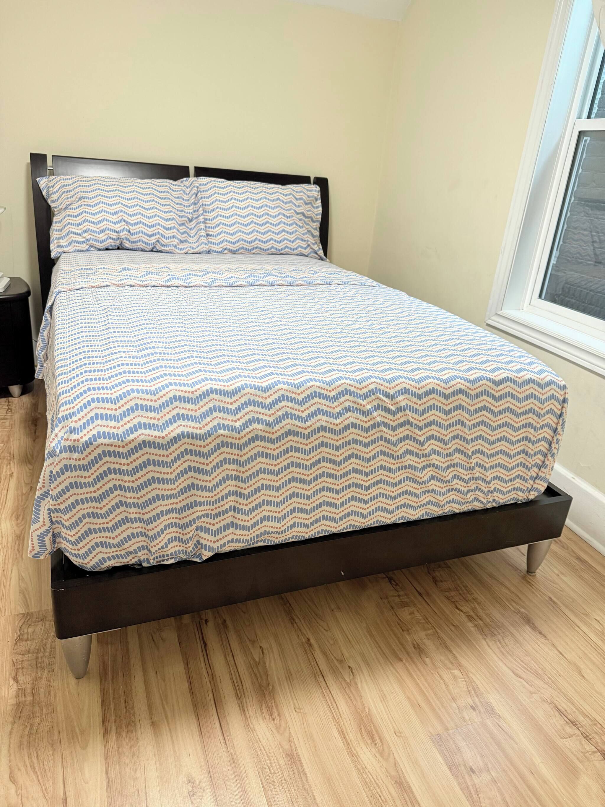 A bed with a patterned bedspread and pillows, a wooden floor, a nightstand, and a window with a view.