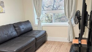 Una sala de estar con un sofá de cuero negro, piso de madera, una ventana con cortinas y un mueble de televisión negro con un control remoto.