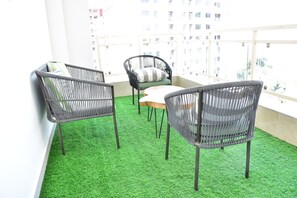 Ein Balkon mit künstlichem Grünrasen, zwei Korbstühle, ein kleiner Tisch und Blick auf hohe Gebäude.