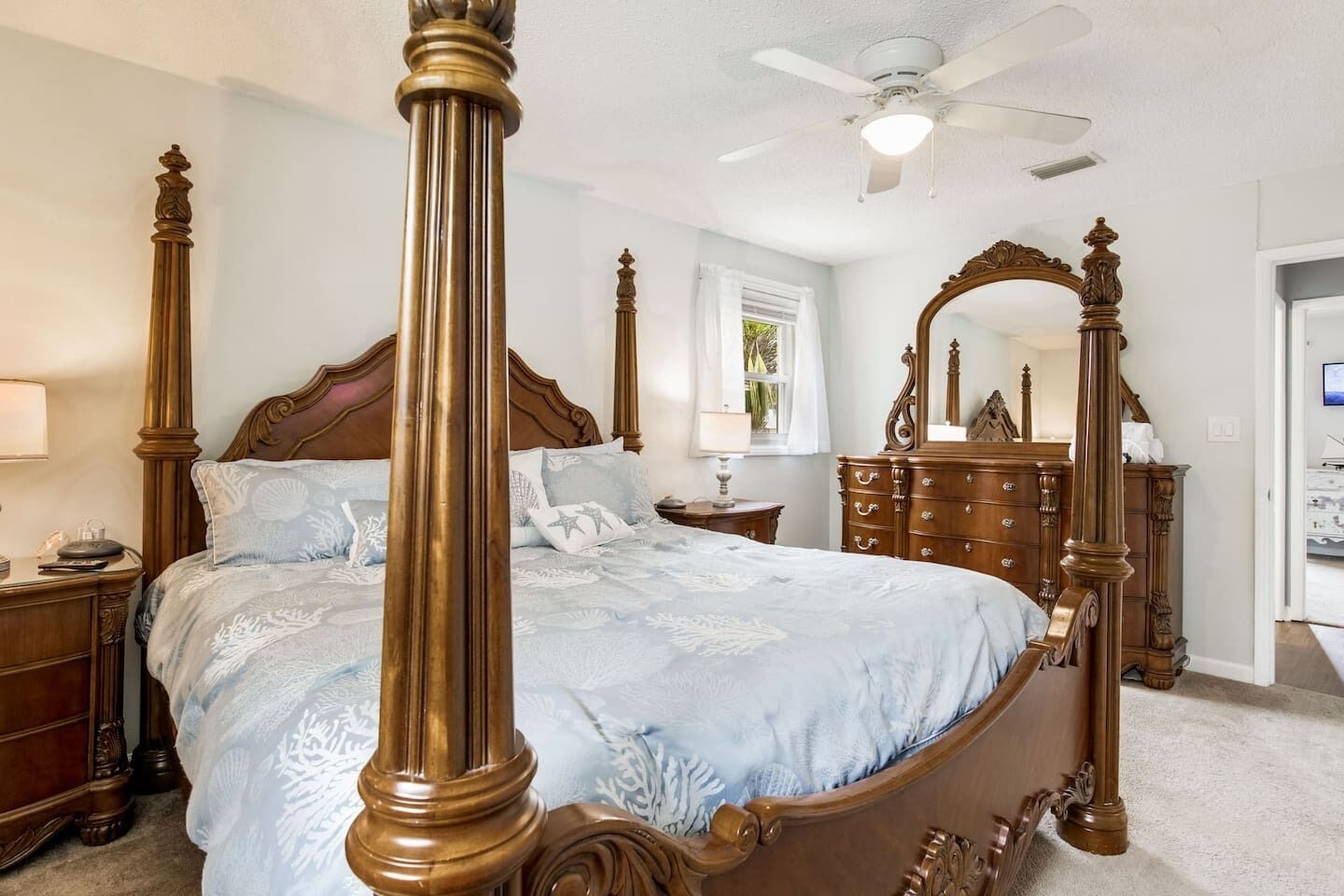 A four-poster bed with a wooden frame and a light blue bedspread. There is a ceiling fan above the bed, and a wooden dresser with a mirror to the right.