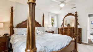 Un lit à baldaquin avec un cadre en bois et une couvre-lit bleu clair. Un ventilateur de plafond est installé au-dessus du lit, et une commode en bois avec un miroir se trouve à droite.