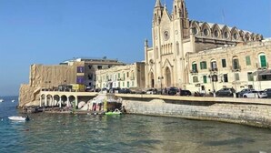 A coastal building with a prominent tower and arches, adjacent to a beach with people and a green boat.