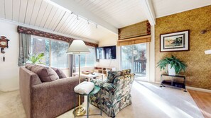 A living room with a brown sofa, a patterned armchair, a floor lamp, and a television. - Bright Rustic Corner Unit, Centrally Located. Private Deck, Summer Pool (Mammoth Lakes)