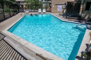 Una piscina rectangular con agua azul cristalina, rodeada por un solárium de concreto. Hay dos sillas reclinables blancas y una reja metálica negra.