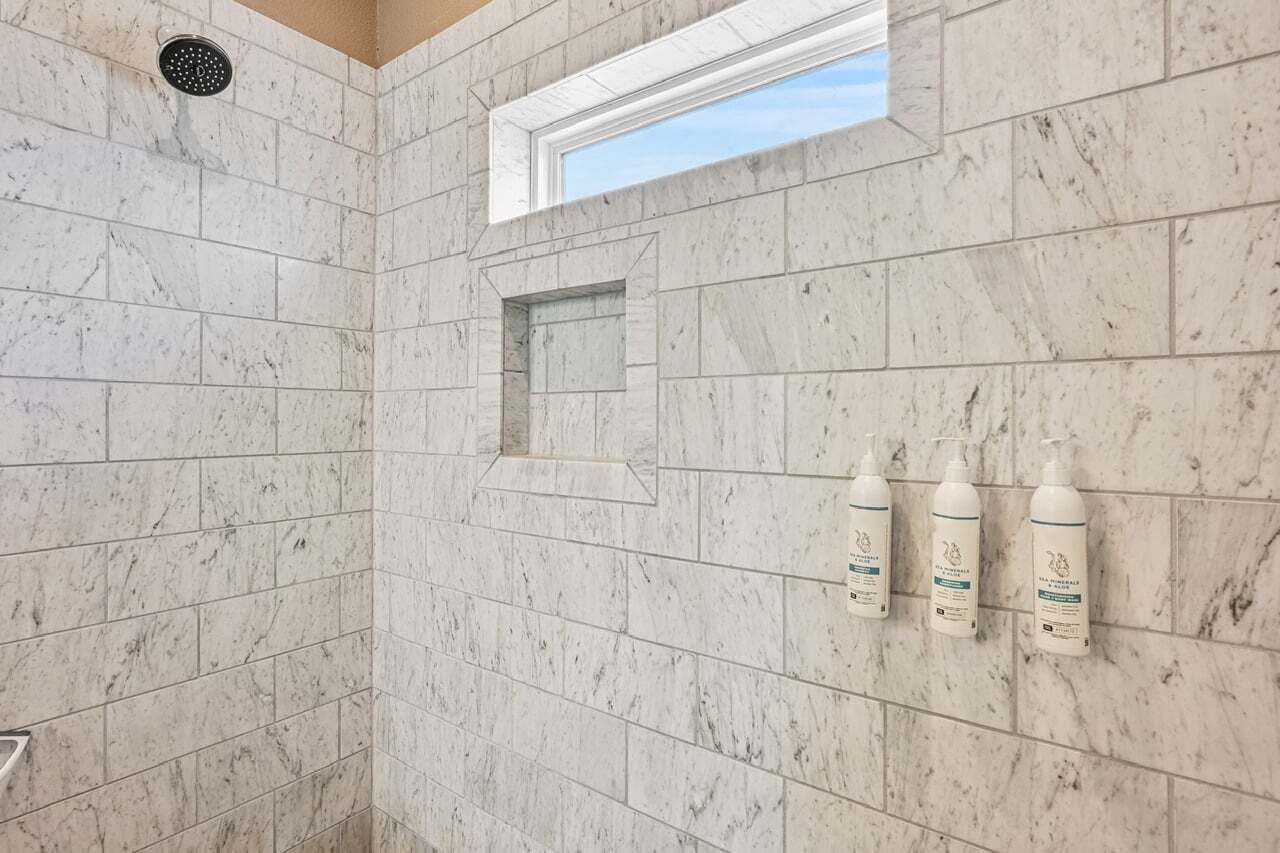 Ein Badezimmer mit Marmorfliesen, einer Duschbrause, einem Fenster und drei Shampoo-Flaschen.