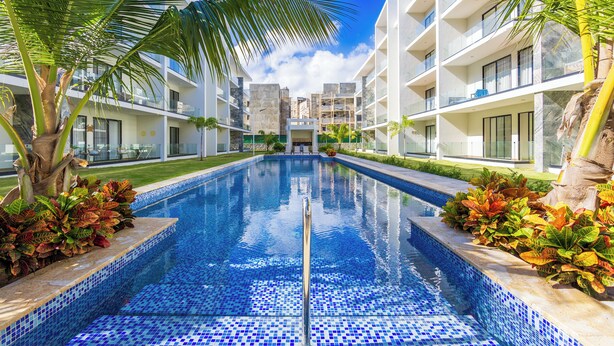 Un hotel moderno con piscina centrale, circondato da palme e un cielo azzurro intenso.